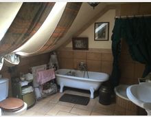 Ein Badezimmer im rustikalen Stil mit Dachschrägen, verzierten Vorhängen und einer freistehenden weißen Badewanne. Es gibt beige Fliesen, einen Waschtisch, einen Toilettenbereich und dekorative Elemente wie Bilderrahmen, eine Milchkanne und Handtuchhalter.