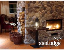 Gemütlicher Innenraum eines Restaurants mit einer Steinwand, die einen modernen Kamin und ein Fach für gestapeltes Brennholz integriert. Vor der Wand stehen ein kleiner Holztisch mit einer Laterne und zwei geflochtene Körbe, daneben ist ein roter Sessel sichtbar.