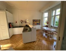 Helle Küche mit integrierter Kochinsel und Esstischbereich in einem Raum mit großen Fenstern und Holzboden. Weiße Einbauküchenmöbel, ein runder Holztisch mit weißen Stühlen, eine farbenfrohe Wandmalerei und dezent eingesetzte Beleuchtung prägen die Gestaltung.