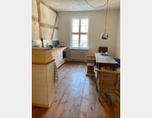 Wohnbereich mit offener Küche in rustikalem Stil. Sichtbare Holzbalken an der Wand, Holzboden, eine schlichte Einbauküche, ein großer Esstisch mit Holzstühlen und Fenster mit weißem Rahmen, das viel Tageslicht hereinlässt.