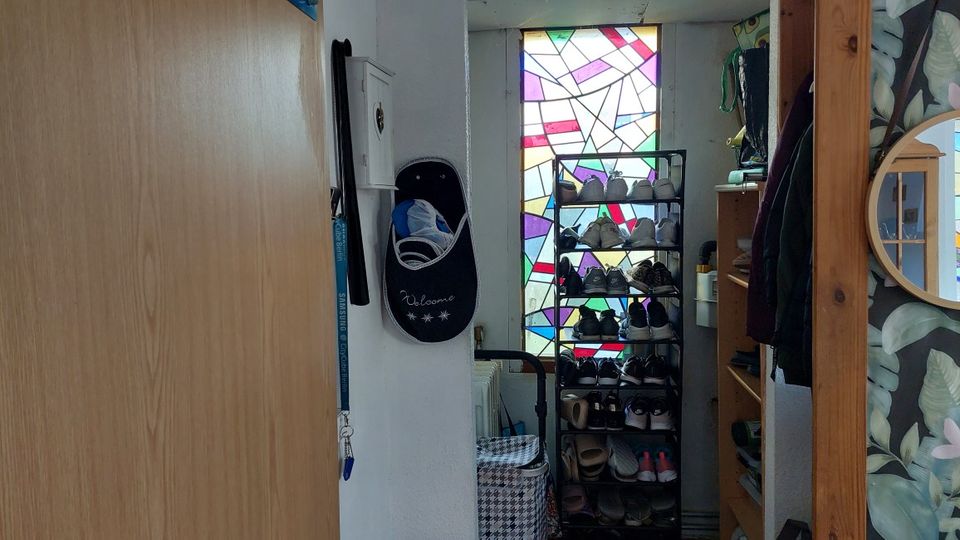 Schmaler Eingangs- bzw. Flurbereich mit einem hohen Schuhregal und Garderobenablage vor einem großen, bunt mosaikartigen Fenster. Links eine helle Holztür und ein Wandhaken, rechts ein offenes Regal und ein runder Spiegel an einer gemusterten Tapete.