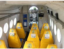 Innenansicht eines kleinen Flugzeugs mit gelben Sitzen, runden Fenstern an den Seitenwänden und Blick auf das Cockpit im Hintergrund. Die Sitze sind mit Logos und Informationsblättern versehen, und die Kabine weist eine schlichte, funktionale Gestaltung auf.