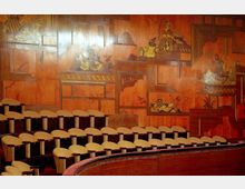 Halbrunder Versammlungsraum mit hölzernen Sitzreihen und einer Wandverkleidung aus poliertem Holz, die mit kunstvoll gestalteten, orientalisch inspirierten Intarsien dekoriert ist. Die Motive zeigen Szenen mit Figuren, Landschaften und geometrischen Elementen.