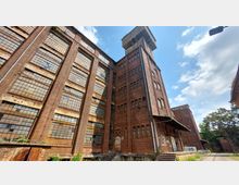 Backstein-Industriegebäude mit hohen, vergitterten Fenstern und einem erhabenen Turm auf der rechten Seite. Das Gebäude zeigt Gebrauchsspuren, und der Bereich ist von einem teils bewachsenen Industriegelände umgeben, unter blauem Himmel mit wenigen Wolken.