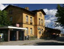 Gelbes Backsteingebäude des Bahnhofs Berlin-Wannsee mit historischen Fenstern und einem kleinen Eingangsbereich mit DB-Logo. Im Vordergrund führt ein gepflasterter Gehweg vorbei, umgeben von Bäumen und Straßenelementen.