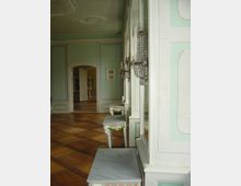 Barocker Innenraum mit pastellgrünen Wänden, verzierten Wandpaneelen und Kristall-Wandleuchtern. Auf dem Holzboden stehen kleine Tische mit marmorierten Oberflächen, und eine offene Tür führt zu weiteren Räumen.