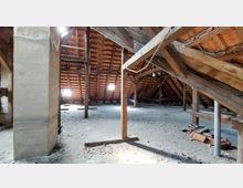 Ein leerer Dachboden mit sichtbarer Holzstruktur und Ziegeldach, gestützt von Balken und Pfeilern. Der Boden ist aus Holz und teilweise mit Schutt bedeckt, Licht fällt durch kleine Fenster.