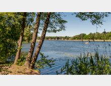 Ein See mit klarem Wasser, umgeben von Bäumen und Schilf am Ufer. Im Hintergrund sind Häuser und ein kleines Boot mit zwei Personen auf dem Wasser zu sehen.
