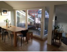 Esszimmer in einer modernen Wohnung mit Holzboden. Ein großer Holztisch mit verschiedenen Stühlen steht vor einer Glasfront, die auf einen Balkon mit Sitzmöbeln und Blick auf Nachbarhäuser führt.