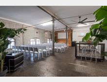 Ein moderner Veranstaltungsraum mit industriellem Stil, gekennzeichnet durch Betonwände, freiliegende Metallpfeiler und große Bogenfenster. Der Raum ist mit weißen Stuhlreihen und einer Leinwand ausgestattet, wobei im Vordergrund Pflanzen und eine kleine Sitz- sowie Stehtischecke sichtbar sind.