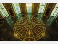 Großer Innenraum mit glänzendem Marmorboden und einem kreisförmigen Muster in der Mitte. Im Hintergrund stehen große Fenster mit Rahmen aus Stein und darüber hängt ein gläserner Kronleuchter.