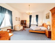 Helles Schlafzimmer mit Doppelbett aus Holz, Nachttischen und Tischlampen sowie einem kleinen Sitzbereich mit Sofa und rundem Beistelltisch. Große Fenster mit dunkelblauen Vorhängen, Kronleuchter an der Decke und klassisches Holzmobiliar wie Schrank und Vitrine prägen den Raum.