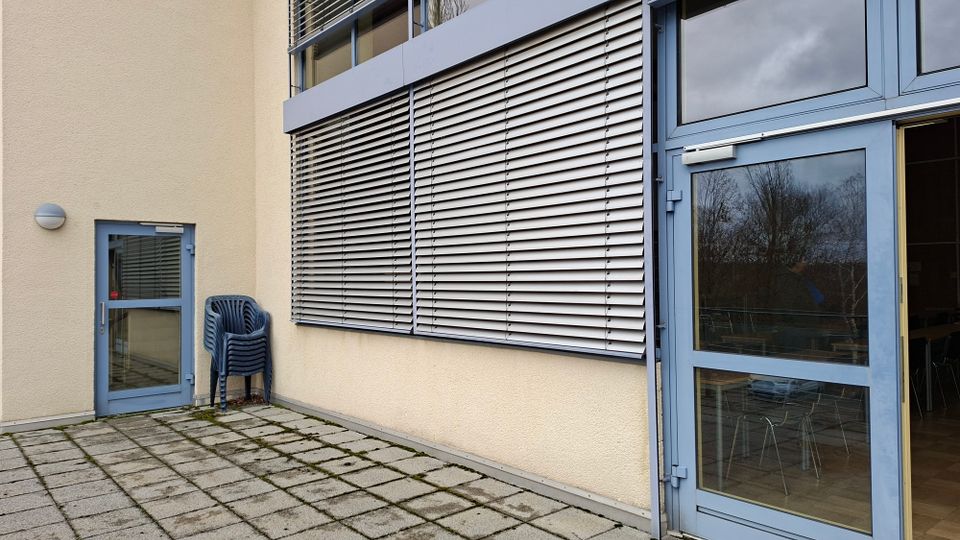 Außenbereich an einem modernen Gebäude mit hell verputzter Fassade, großen Fenstern mit horizontalen Außenjalousien und einer blau gerahmten Glastür. Der Boden ist mit quadratischen Betonplatten gepflastert; neben einer kleineren Nebentür stehen gestapelte Kunststoffstühle.