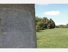 Steinsäule mit eingraviertem Text im Vordergrund auf einer grünen Wiese, umgeben von Bäumen und unter einem klaren blauen Himmel. Der Text erläutert historische Informationen zu dem Park, der im Hintergrund sichtbar ist.