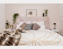 Ein modernes Schlafzimmer mit einem großen Bett, das weiße und gemusterte Kissen sowie eine goldgemusterte Tagesdecke zeigt. Der Hintergrund besteht aus einem rosa Kopfteil mit Marmoroptik, eingerahmt von Nachttischen mit Pflanzen und minimalistischen Lampen.