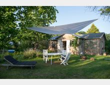 Steingebautes Gartenhaus mit geöffneten weißen Fensterläden, umgeben von Bäumen und Pflanzen. Im Vordergrund Gartenmöbel und ein Sonnensegel über einer grünen Wiese.