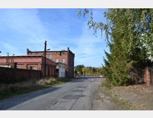Straßenszene mit Kopfsteinpflaster und Schienen, flankiert von einem Backsteingebäude mit roten und weißen Fassaden sowie Bäumen rechts im Bild. Im Hintergrund sind weitere Vegetation und ein parkendes Auto zu sehen.