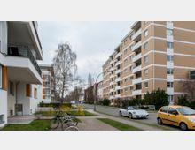 Wohnsiedlung mit mehrstöckigen, hellbraun verklinkerten Wohngebäuden und Balkonen; im Vordergrund eine grüne Rasenfläche mit Fahrradständern und geparkten Autos entlang der angrenzenden Straße. Kahle Bäume und Sträucher säumen den Gehweg.