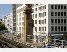 Stadtansicht mit erhöhten Bahnschienen im Vordergrund, einer historischen Steinsäule mit Reliefverzierungen sowie modernen Gebäuden im Hintergrund, darunter eine Bankfiliale im Erdgeschoss eines der Gebäude. Links im Bild sind Bäume zu sehen.