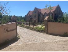 Ein gepflasterter Weg führt durch ein Tor mit niedrigen Mauern und der Aufschrift „Rosencarrée“ zu einem großen, mehrstöckigen Gebäude aus Backstein mit Giebelarchitektur und roten Ziegeldächern. Im Vordergrund sind Blumenbeete, ein Holzgeländer und vereinzelte Bäume zu sehen.