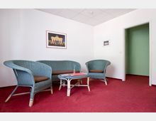 Wartebereich mit roten Teppichboden, beigen Wänden und drei türkisfarbenen Korbmöbeln mit braunen Sitzpolstern. Ein niedriger Glastisch steht in der Mitte, an der Wand hängt ein gerahmtes Bild des Brandenburger Tors.