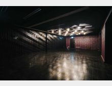 Ein dunkler, leerer Raum mit glänzendem schwarzem Boden, einer gemusterten Wand aus vertikalen Streben auf der linken Seite und einer violetten Wand im Hintergrund. Von der Decke hängen mehrere runde Lampen, deren Licht Schattenmuster auf den Boden und die Wände wirft.