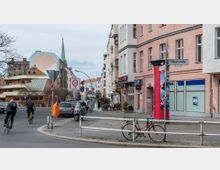 Straßenszene mit mehrstöckigen Wohn- und Geschäftsgebäuden, darunter ein pastellrosa Gebäude mit Schildern wie "Hauptstraße" und "Café Sky". Im Vordergrund geparkte Fahrräder, Autos, Verkehrsschilder und zwei Radfahrer, und im Hintergrund ein modernes Gebäude mit auffälligem grünem Dach und ein Kirchturm.