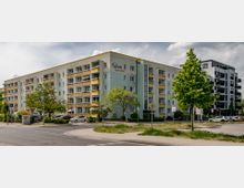 Mehrfamilienwohnhaus mit fünf Etagen, gelben Balkonen und grünlicher Fassade, umgeben von Bäumen und parkenden Autos; daneben ein moderner, schwarzer Neubau. Im Vordergrund befindet sich eine Straßenkreuzung mit Gehwegen und Verkehrsschildern.