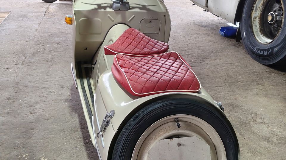 Überdachter Industriehof oder Lagerhalle mit Betonboden und Stützpfeiler, davor eine offene Rasenfläche und alte Backsteingebäude. Im Vordergrund steht ein Motorroller mit Beiwagen, daneben ist die Seite eines großen Busses sichtbar.