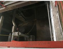 Blick in einen technischen Schacht mit Rohren, Ventilen und einer rostfarbenen Metallkonstruktion am Rand. Die Wände und die Umgebung des Schachts wirken roh und unbehandelt.