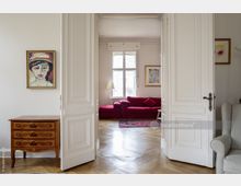 Helle Wohnzimmerszene mit Doppelflügeltür in klassischem Weiß, einem Parkettboden und roten Sofas vor einem großen Fenster. Links ein Holzkommode mit Intarsien und einem expressionistischen Porträt an der Wand, im Hintergrund ein bunter Teppich und weitere Kunstwerke.