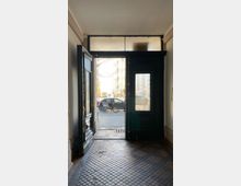 Blick aus einem Hauseingang mit einer geöffneten, dunkel gestrichenen Holztür und Glaseinsatz. Der Boden ist mit kleinen Fliesen gestaltet, und im Hintergrund sind eine Straße, parkende Fahrräder und Autos zu sehen.