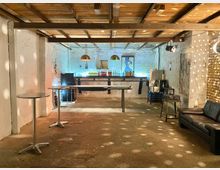 Rustikaler Partyraum mit Holzdecke, unverputzten Wänden und Backsteinelementen. Im Vordergrund stehen Stehtische, im Hintergrund eine Bar mit Flaschen und Hängelampen; punktuelle Beleuchtung erzeugt Lichtreflexe auf den Wänden.