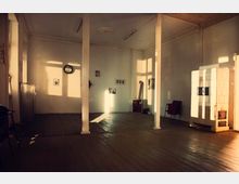 Ein spärlich eingerichteter Raum mit Holzdielenboden und weiß gestrichenen Wänden, durchbrochen von zwei weißen Säulen. An der Wand hängen kleine Bilder und eine Wanduhr, während ein Kachelofen und ein schlichter Metallofen an der rechten Seite stehen.