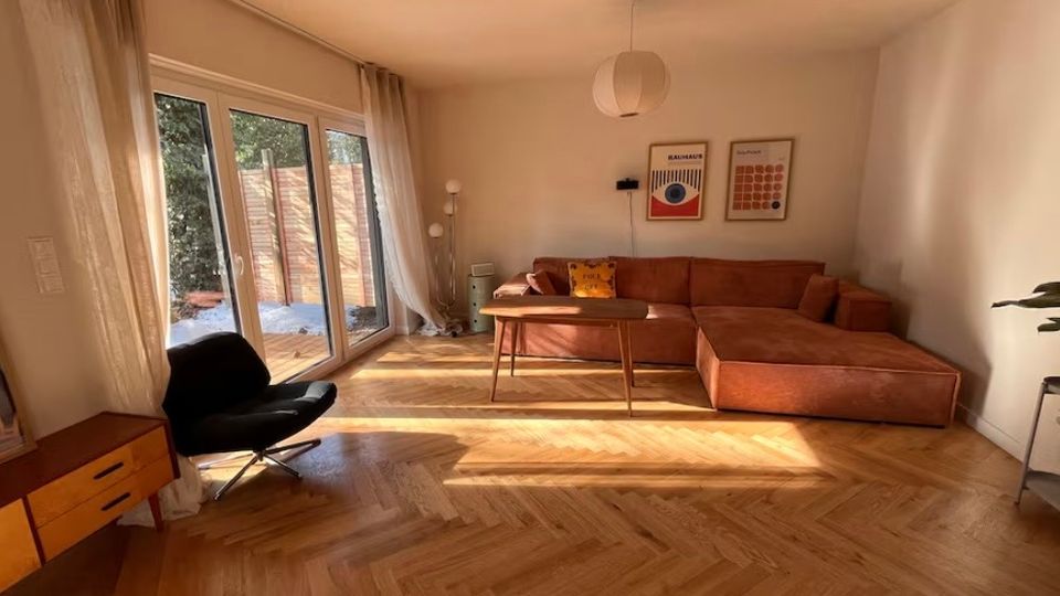 Wohnzimmer mit hellem Fischgrätparkett, einer großen orangefarbenen Eckcouch, einem kleinen Holztisch und einem schwarzen Sessel. Links befindet sich eine große Glastür mit cremefarbenen Vorhängen, die zu einer Terrasse führt, während zwei gerahmte Bilder an der beigen Wand hängen.