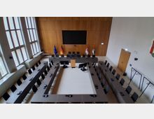 Moderner Sitzungssaal mit U-förmig angeordneten Tischen und zahlreichen Stühlen, ausgestattet mit Mikrofonen und Monitoren. Vor einer holzverkleideten Wand hängen ein großer Bildschirm und mehrere Flaggen; links sorgen hohe Fensterfronten für Tageslicht.