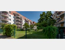 Mehrstöckiges Wohngebäude mit roten und weißen Fassaden und Balkonen, das von einem eingezäunten Grünbereich mit Bäumen und Sträuchern umgeben ist; im Vordergrund ein Metallzaun mit Efeubewuchs.