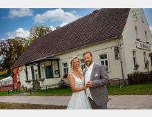 Ein Hochzeitspaar steht vor einem historischen Gebäude mit heller Fassade, grünen Fensterrahmen und einem dunkelroten Ziegeldach. Neben dem Haus befinden sich Blumenkästen, ein kleiner Gartenzaun und eine befestigte Gehwegfläche.