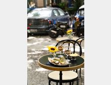 Ein kleiner runder Tisch im Freien mit schwarzer Platte und goldener Umrandung, darauf zwei Gläser Roséwein, ein Teller mit Brot und Beilagen, eine Dose mit Speisen und eine Vase mit gelber Sonnenblume; im Hintergrund geparkte Autos und ein motorisiertes Fahrrad.