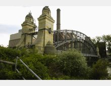 Historische Brücke mit steinernen Pfeilern und schmiedeeisernen Verzierungen, flankiert von bewachsener Vegetation; im Hintergrund sind Backsteinbauten, ein Schornstein und moderne Architektur sichtbar.