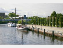 Uferpromenade mit Anlegestellen, an denen zwei Ausflugsschiffe liegen, darunter ein Dampfschiff mit rauchendem Schornstein. Im Hintergrund eine Brücke mit Graffiti, daneben historische Säulen und eine Reihe von Bäumen entlang des Gehwegs.