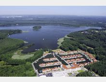 Luftaufnahme einer großen Seenlandschaft mit umliegenden Wäldern und einer kleinen Insel im Wasser. Im Vordergrund eine Ferienanlage mit roten Dächern, angeordnet um künstliche Kanäle und eine Marina.