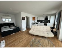 Modernes Apartment mit offener Raumaufteilung, bestehend aus einem Wohn- und Schlafbereich mit einem schwarzen Bett und Wandspiegel, sowie einer kleinen Küche mit weißen Schränken und schwarzen Arbeitsflächen. Der Boden ist aus Holzoptik, es gibt ein beiges Sofa und zwei runde, gläserne Couchtische.