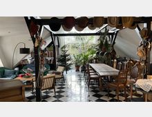 Wohnraum unter einem Dachgeschoss mit Holzbalken und Schrägen. Ein langer Holztisch mit Stühlen steht auf einem schwarz-weiß gefliesten Boden, umgeben von Pflanzen, Regalen und einer Sitzecke mit Sofa und Sesseln; Hüte hängen an den Balken.