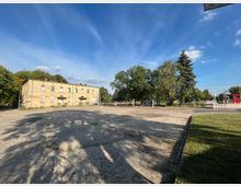 Ein altes, zweistöckiges Gebäude mit verblassener Fassade steht auf einem gepflasterten Platz, umgeben von Bäumen und Grünflächen. Im Hintergrund sind eine Straße, Absperrungen und ein paar Verkehrs- und Bahnanlagen zu sehen, bei klarem Himmel und Sonnenschein.