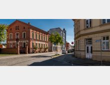 Straßenszene in einer Kleinstadt mit historischen Gebäuden; links ein zweigeschossiges Ziegelhaus mit grünen Fensterläden und der Aufschrift „Bankverein Wittenberge“, rechts ein hellgelbes Eckgebäude mit schmuckvollen Fassadendetails. Im Hintergrund sind weitere Gebäude und eine Apotheke erkennbar.