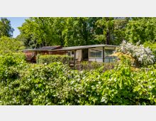 Ein kleines Gartenhaus mit weißer und roter Fassade umgeben von dichter Vegetation, darunter eine Hecke im Vordergrund und blühende Sträucher. Im Hintergrund sind große Bäume unter einem klaren Himmel zu sehen.