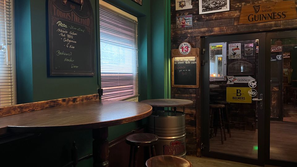 Gemütliche Pub-Ecke mit hohen Holztischen und Barhockern neben einem Fenster mit Jalousien. Dunkelgrüne Wände und eine rustikale Holzvertäfelung mit Schiefertafeln, gerahmten Bildern und Guinness-Werbeschildern sowie einer Glastür zum Nebenraum.