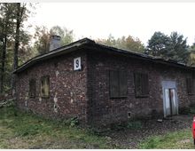 Backsteingebäude mit flachem Dach und mehreren vergitterten oder verschlossenen Fenstern, auf denen Spuren von Verfall sichtbar sind. Die Umgebung besteht aus einer bewaldeten Landschaft mit Bäumen im Hintergrund.
