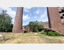 Ehemaliger Fabrikhof mit Backsteingebäuden, einem hohen Schornstein und teils verfallenen Strukturen; im Hintergrund Bäume und blauer Himmel.